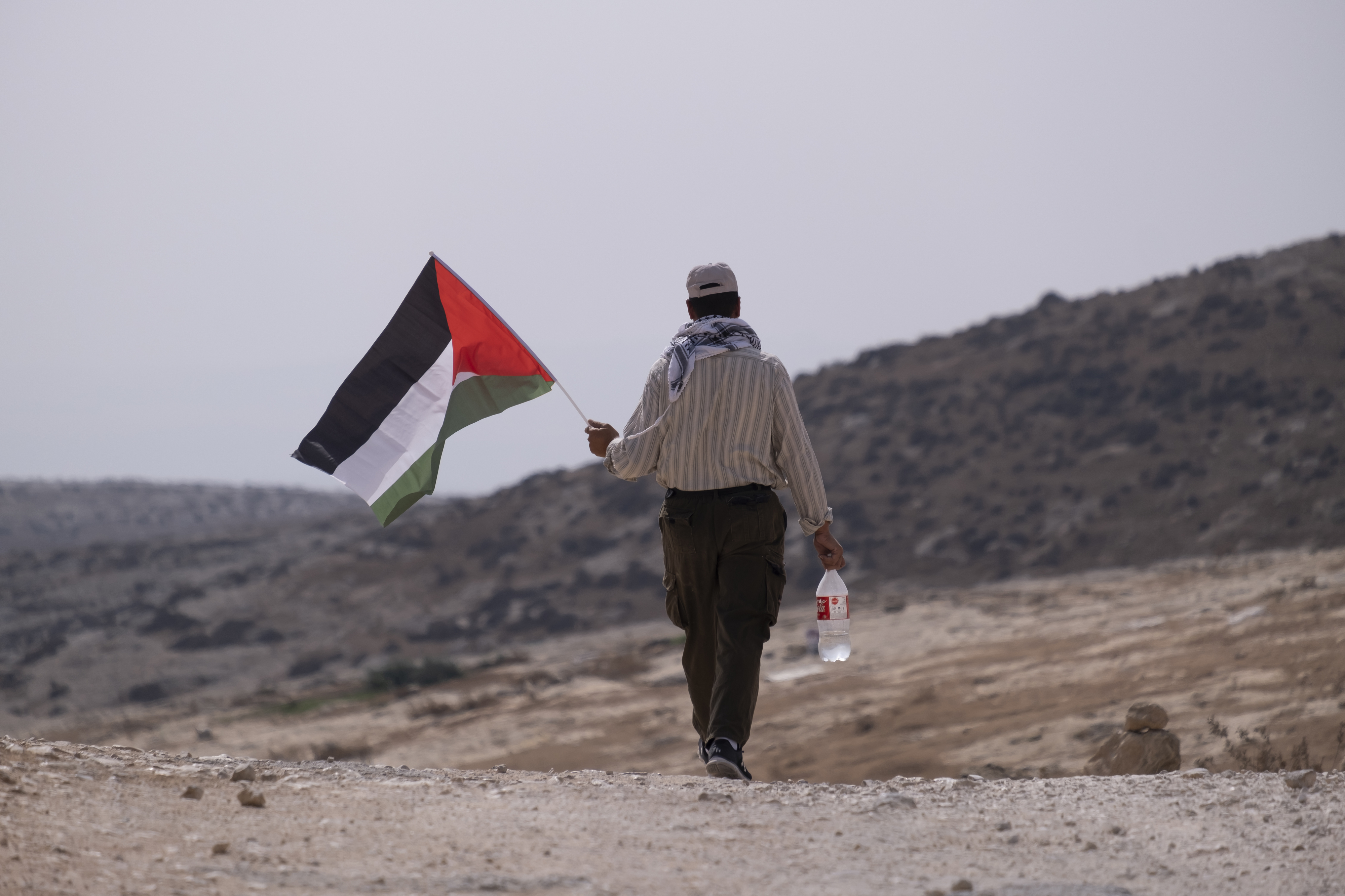 A person is walking away on a rocky terrain, holding a Palestinian flag and a bottle of water. Desolate landscape in the background