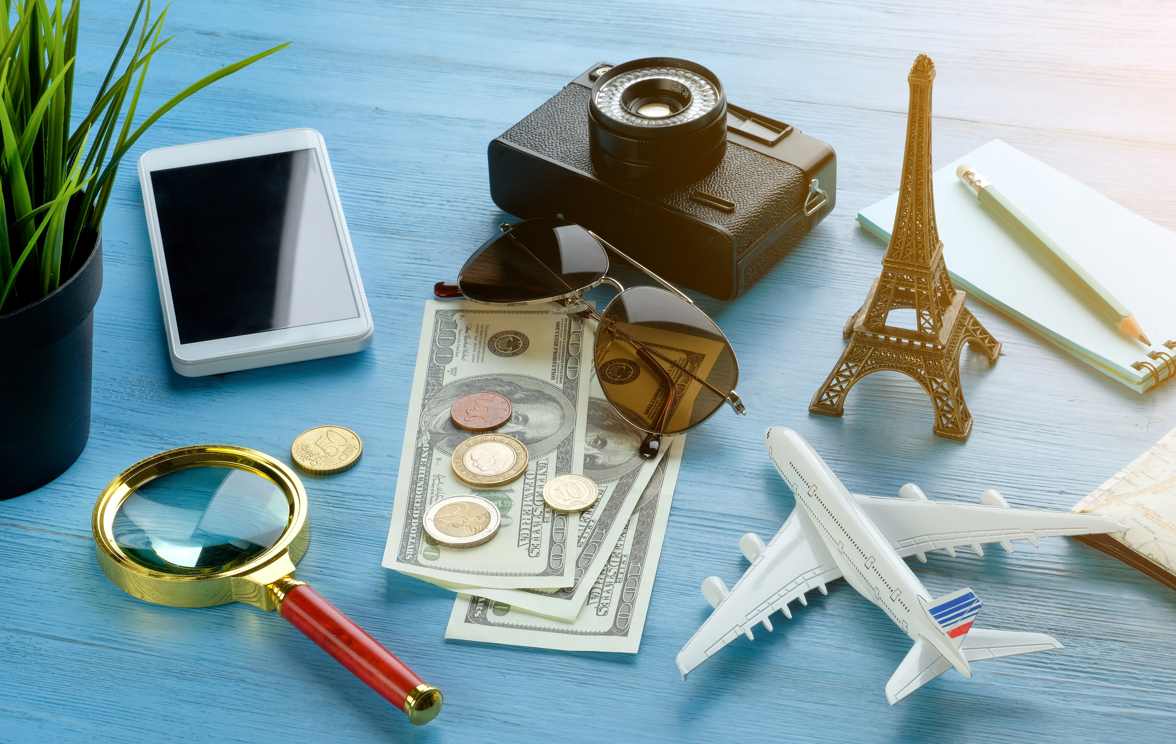 A table with a camera, smartphone, toy airplane, Eiffel Tower model, magnifying glass, plant, sunglasses, notepad, and various currencies
