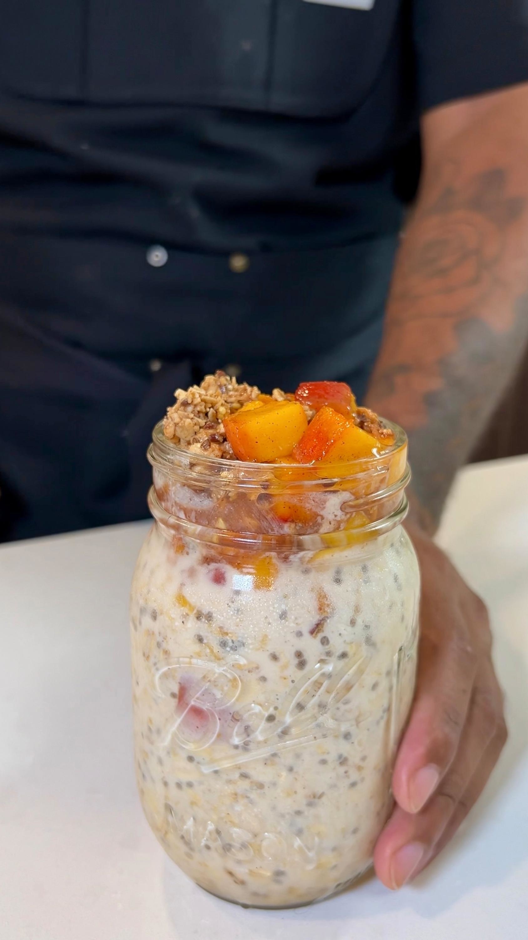 A person holds a mason jar filled with overnight oats, topped with chunks of peach and granola