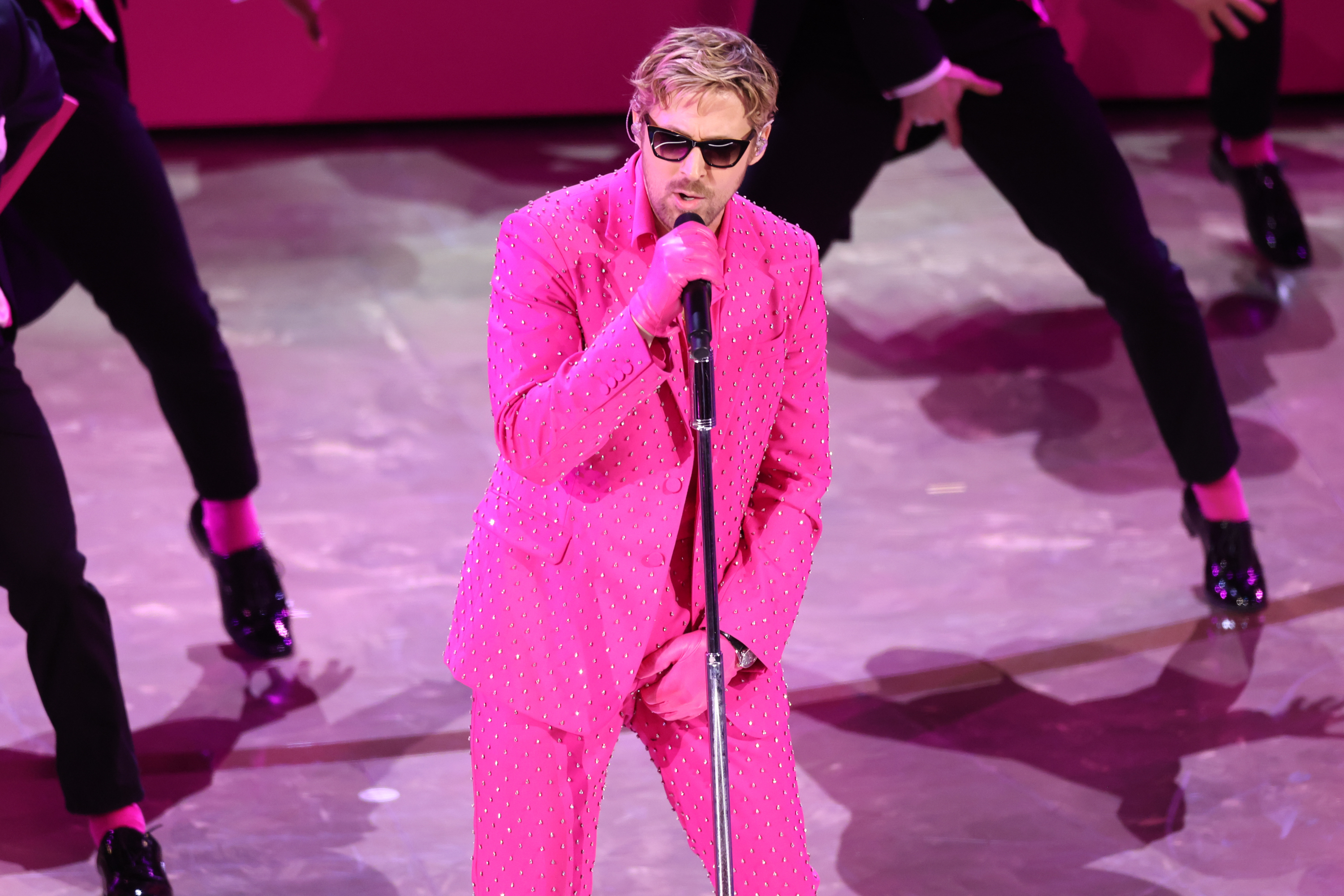 Ryan Gosling wearing a sparkling Barbie pink suit and sunglasses, performing on stage with backup dancers