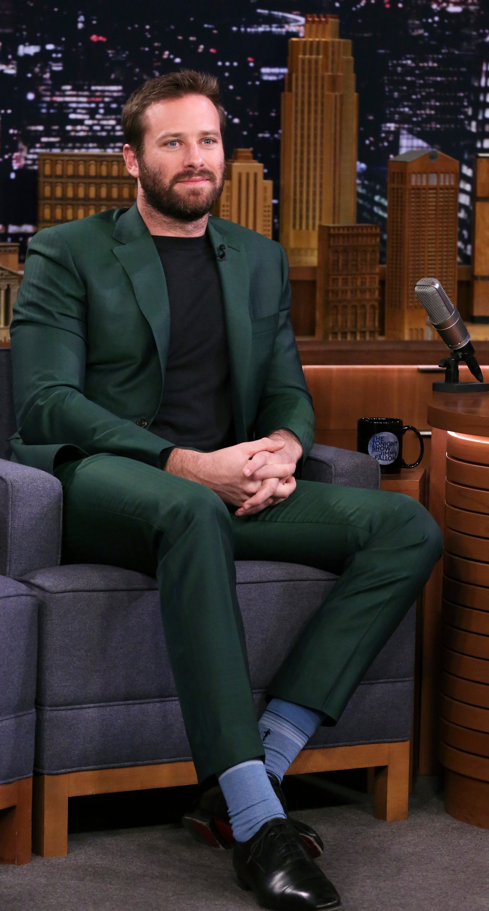 Armie Hammer, wearing a tailored suit with a dark shirt and socks, sits on a talk show set in front of a cityscape backdrop