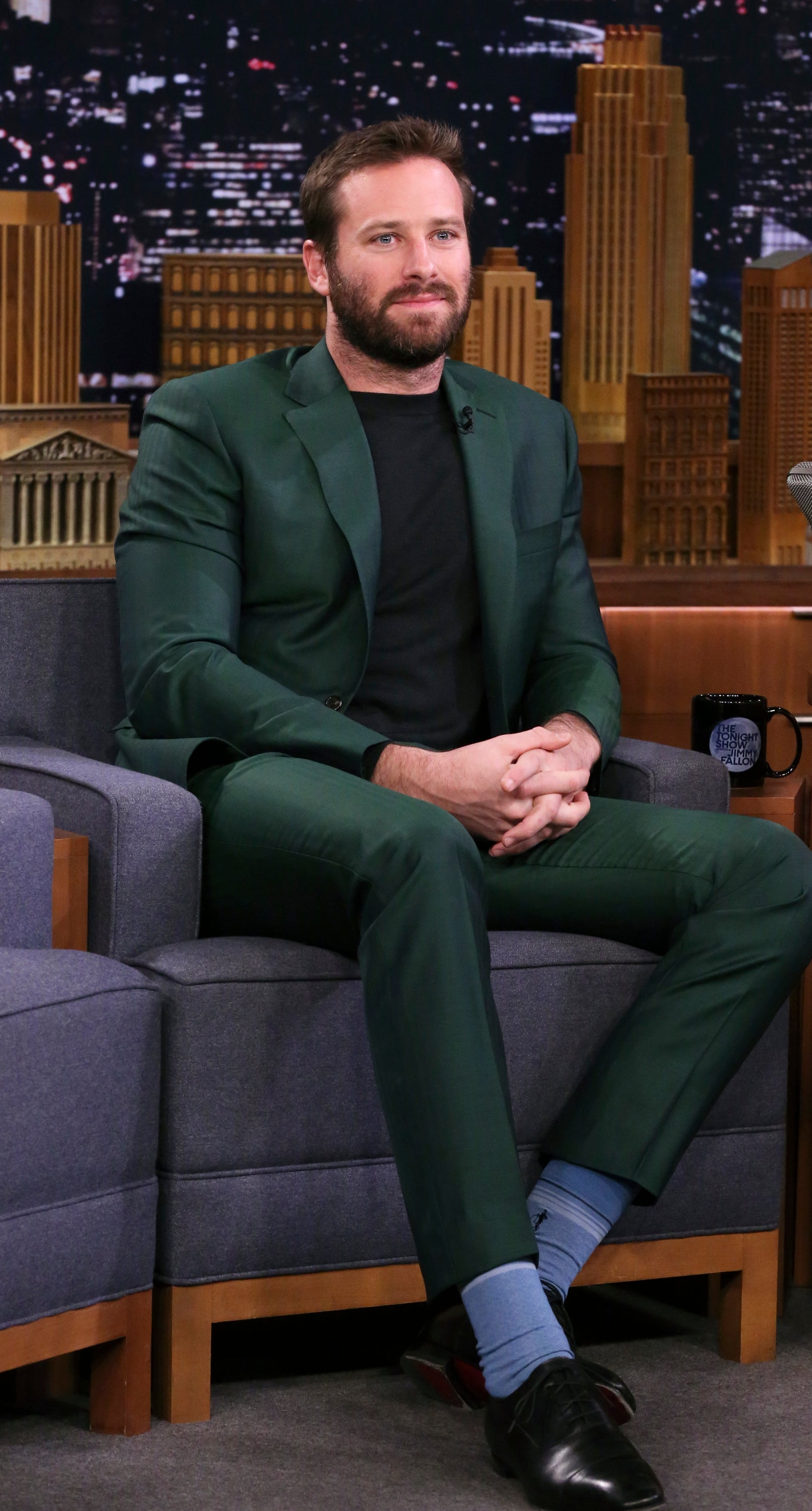 Armie Hammer, wearing a tailored suit with a dark shirt and socks, sits on a talk show set in front of a cityscape backdrop