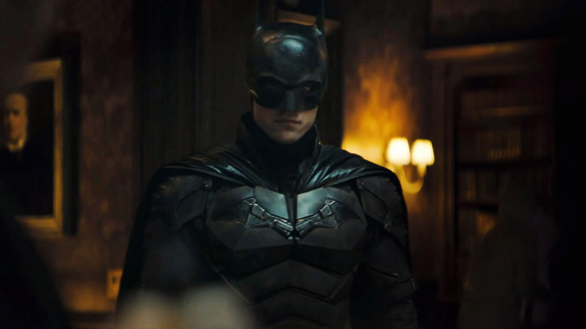 Robert Pattinson dressed as Batman in an armored bat-suit, standing in a dimly lit room with bookshelves and wall sconces in the background