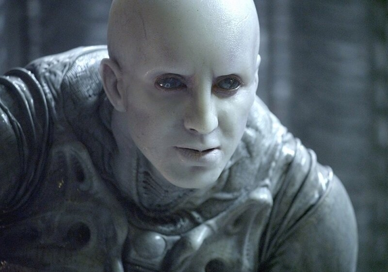 A character from the movie "Prometheus" with pale skin and a bald head wearing futuristic armor in a dimly lit environment