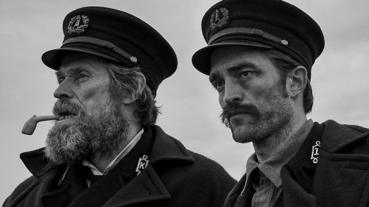Willem Dafoe, smoking a pipe, and Robert Pattinson, both wearing naval uniforms with caps, staring intensely in the same direction