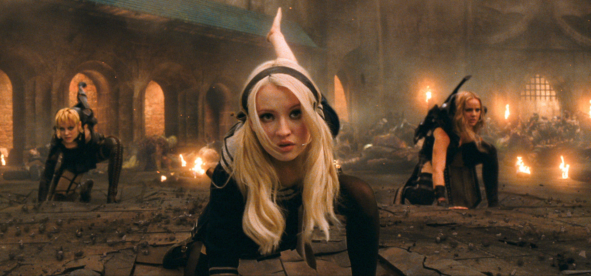Emily Browning, Vanessa Hudgens, and Abbie Cornish in action scene from "Sucker Punch," wearing fantasy-styled combat outfits, crawling forward on a damaged ground