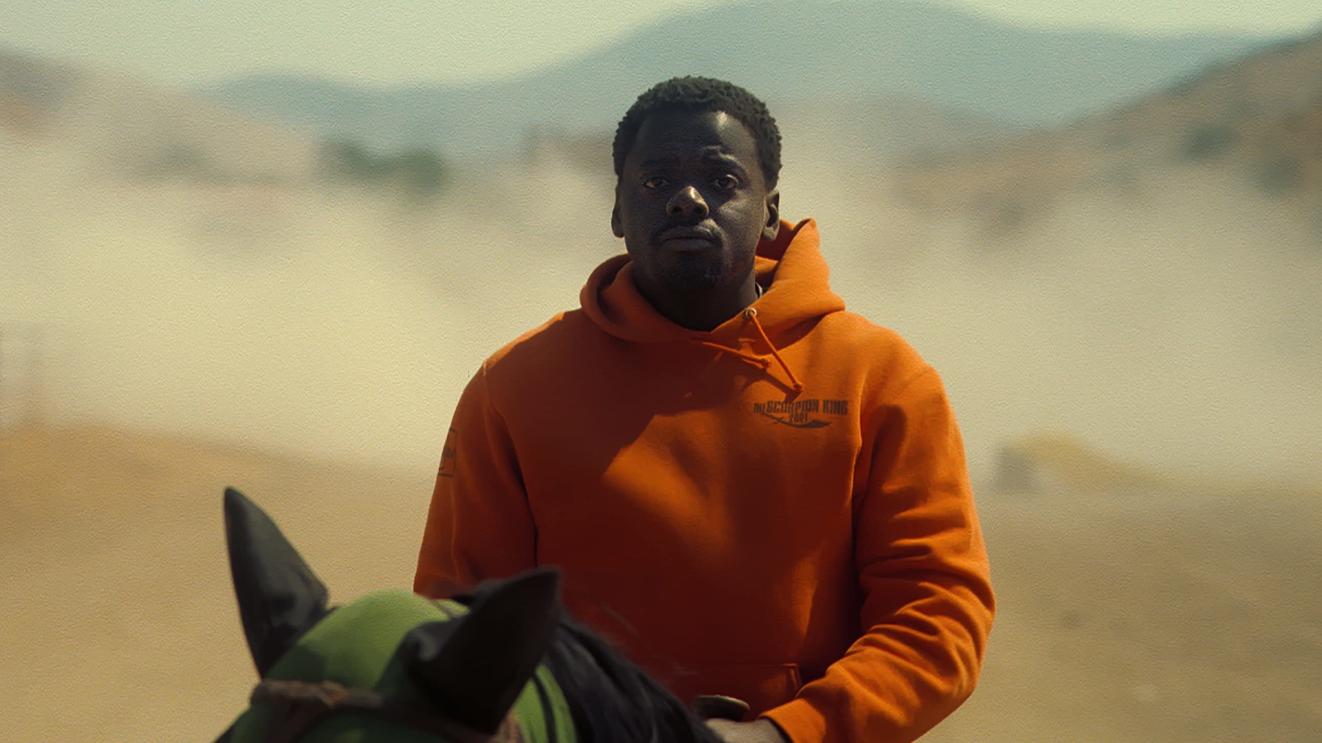 Daniel Kaluuya rides a horse while wearing a casual hoodie. Dust and mountains are visible in the background