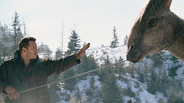 Chris Pratt in a snowy landscape, extending his hand to touch the snout of a dinosaur-like creature on a leash