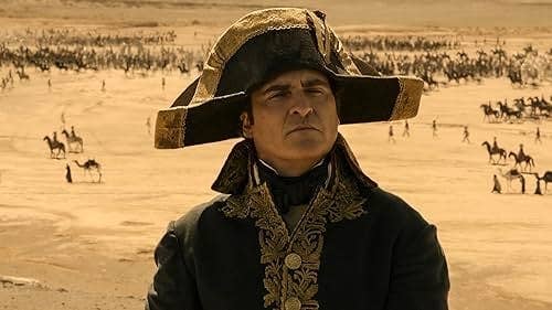 Joaquin Phoenix as Napoleon Bonaparte in period military attire with a distinctive bicorne hat, set against a desert battlefield with soldiers in the background