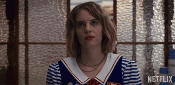 Maya Hawke, wearing a sailor-themed uniform with stripes and a red name tag, stands in front of a glass window