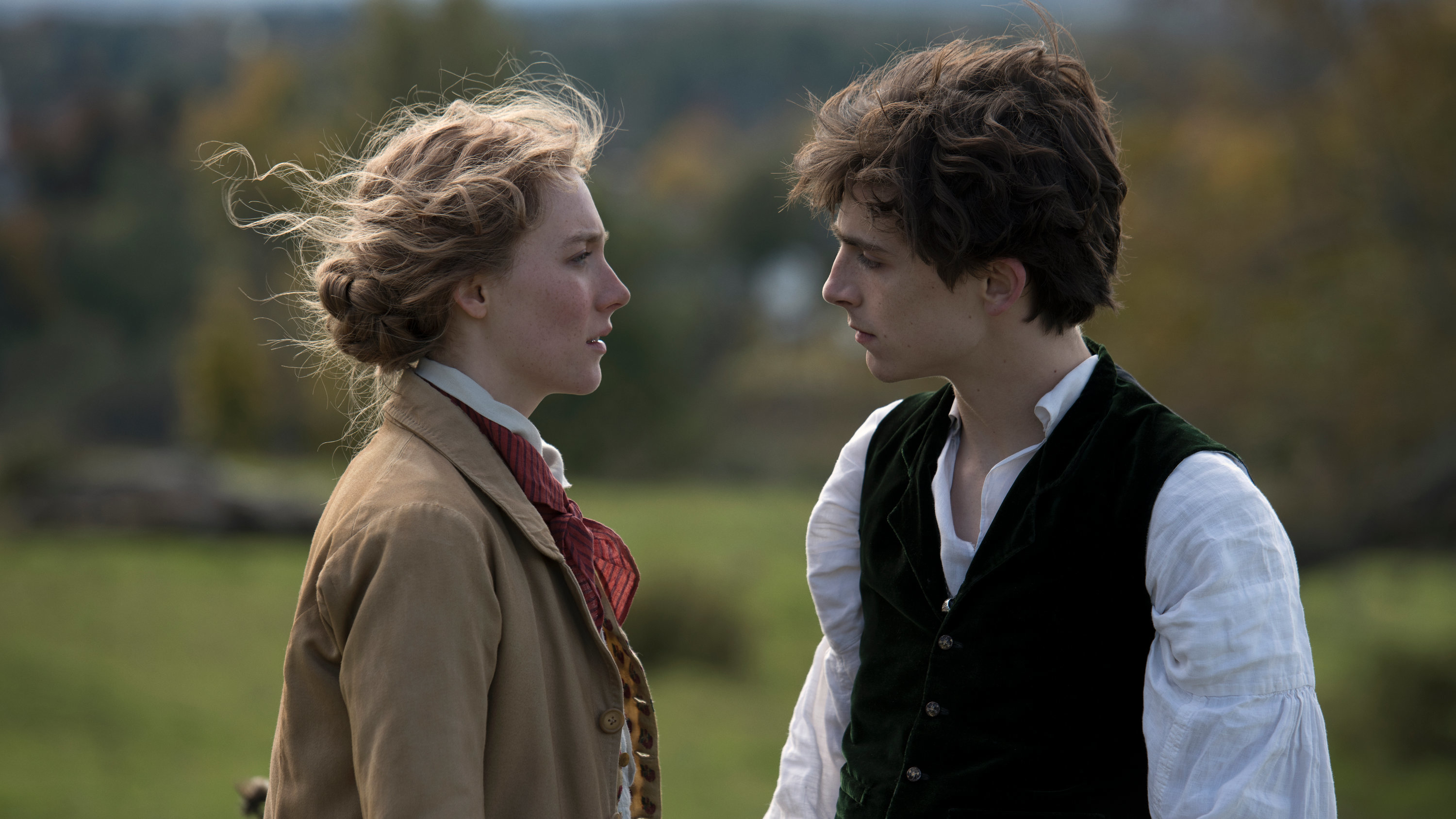 Saoirse Ronan and Timothée Chalamet, dressed in period clothing, gaze intently at each other in a rural outdoor setting