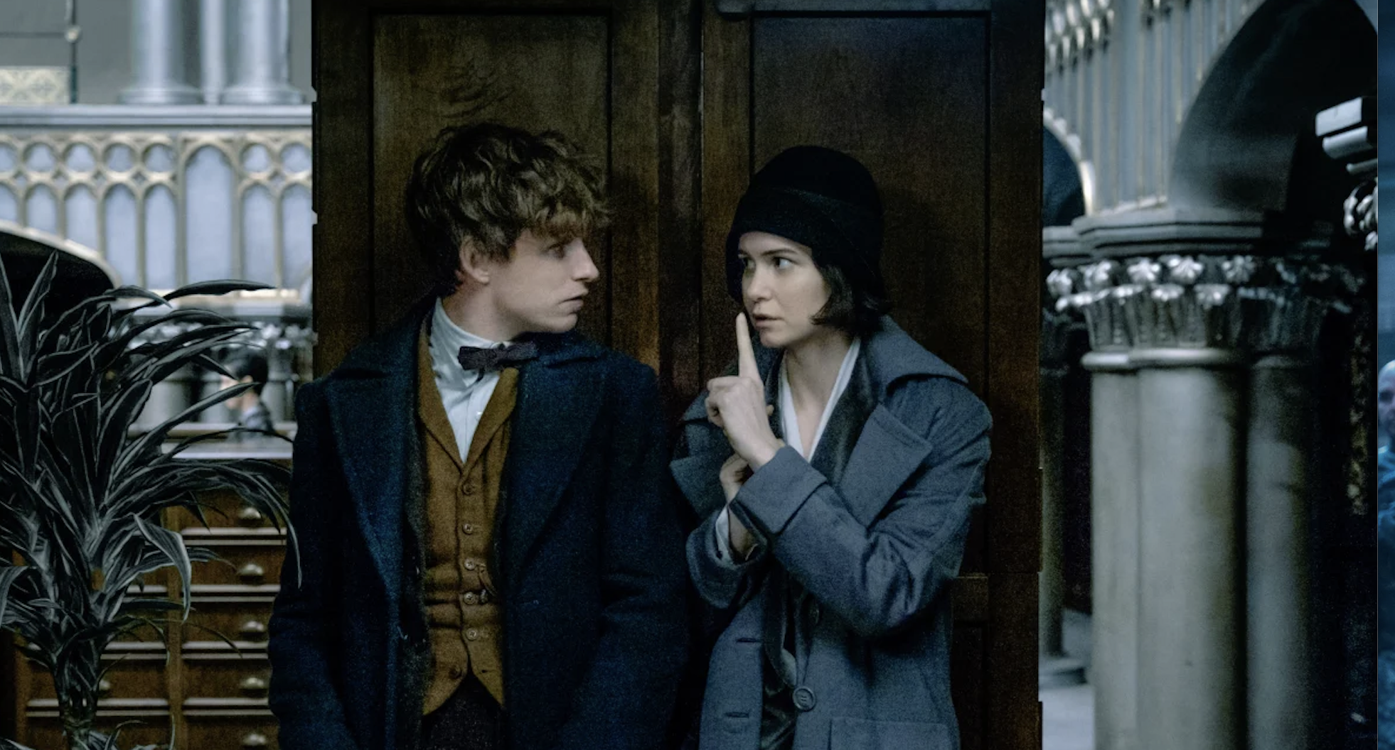 Eddie Redmayne and Katherine Waterston in period attire, standing close to each other in an ornate building, with Waterston's character signaling to stay quiet