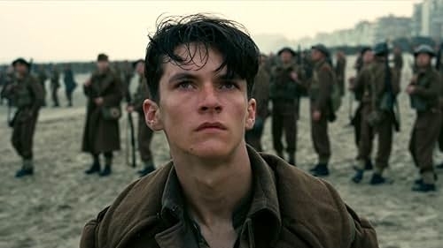 A young man with short dark hair stands on a beach, looking up intently. Soldiers in brown uniforms are behind him