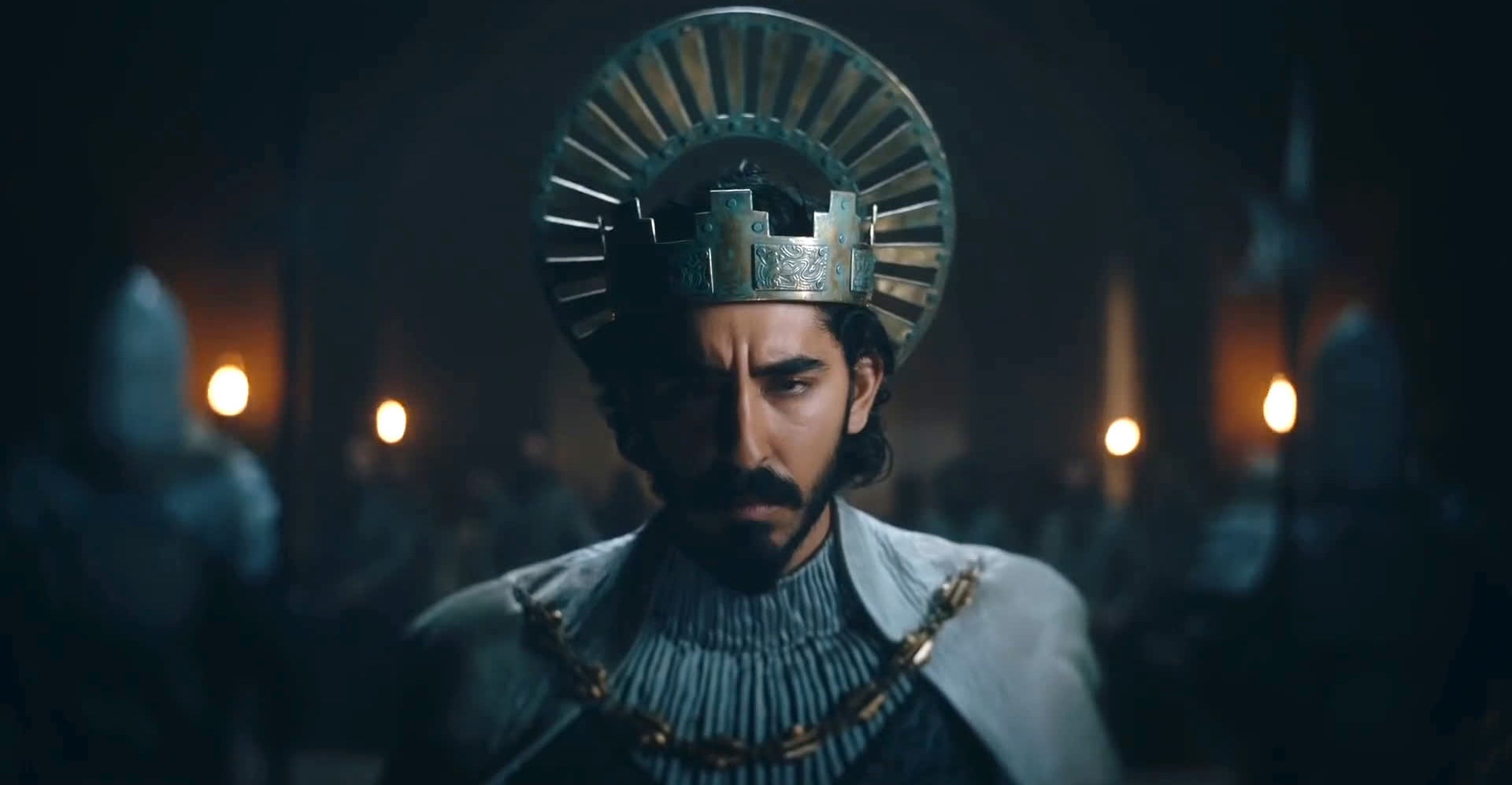 Dev Patel dressed as King Arthur, wearing a metallic crown and armor, in a scene from the movie "The Green Knight."
