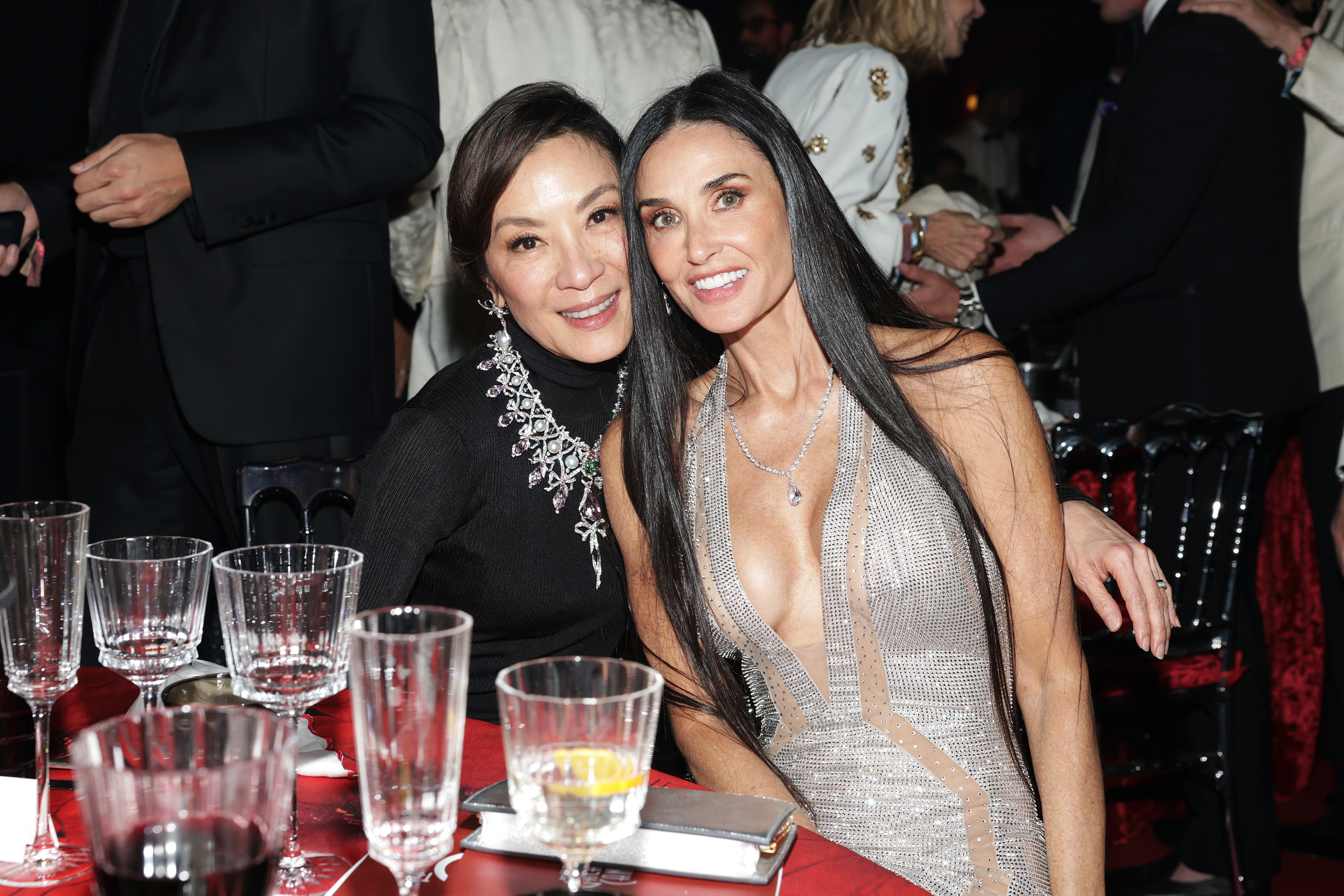 Michelle Yeoh and Demi Moore pose together at a formal event, sitting at a table set with empty glasses