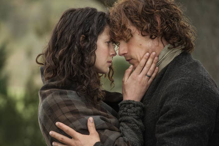 Caitriona Balfe and Sam Heughan, in character, embrace closely with foreheads touching and hands caressing each other's faces in a tender moment