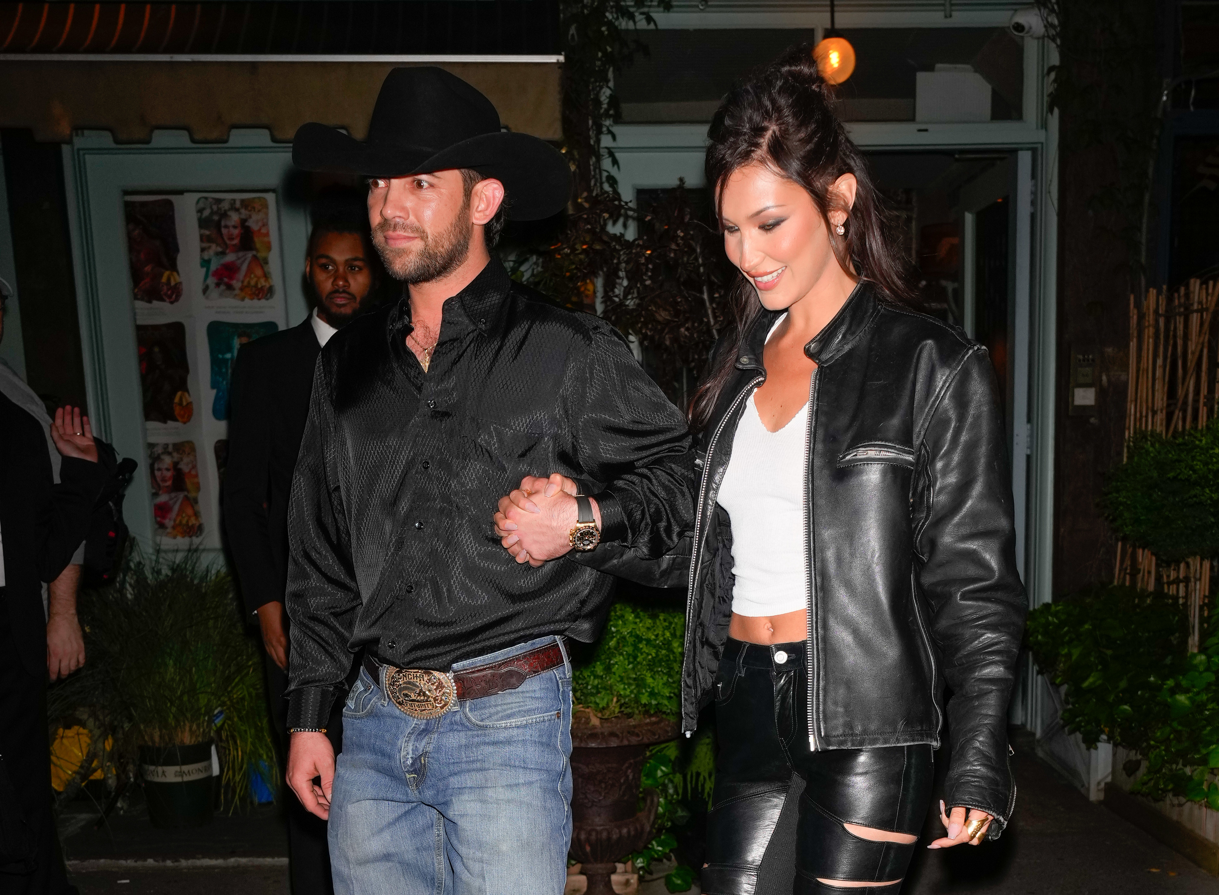 Bella Hadid and Adan hold hands, walking outdoors. Bella wears a top, leather jacket, and ripped pants