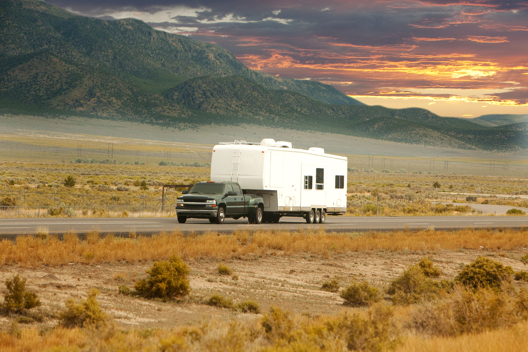 A truck is towing a large white camper on an open road through a desert landscape with mountains in the background during sunset