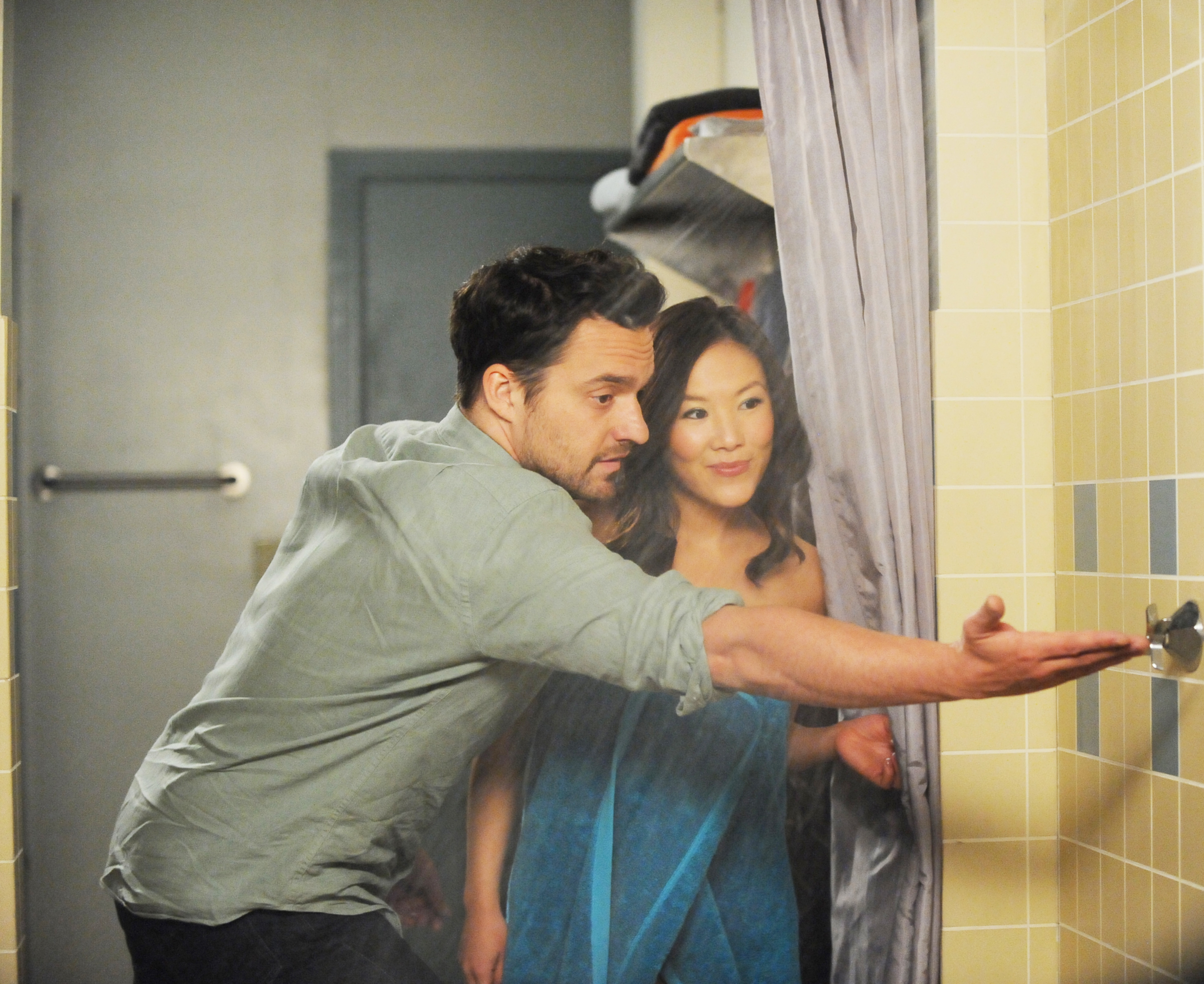 Jake Johnson and Jameela Jamil stand close together in a shower stall, with Jake reaching to turn on the water