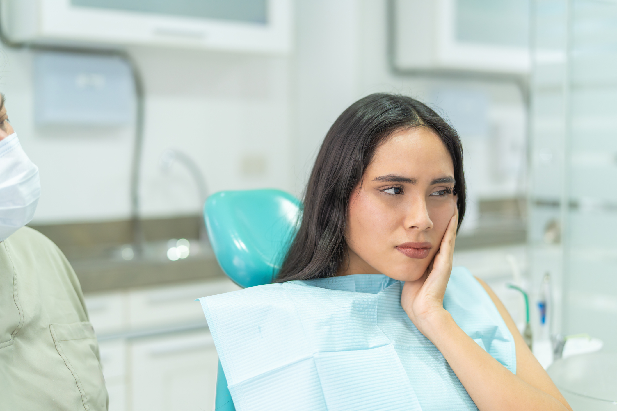 A woman is sitting in a dental chair, holding her cheek with a pained expression. A dental professional in scrubs is visible beside her