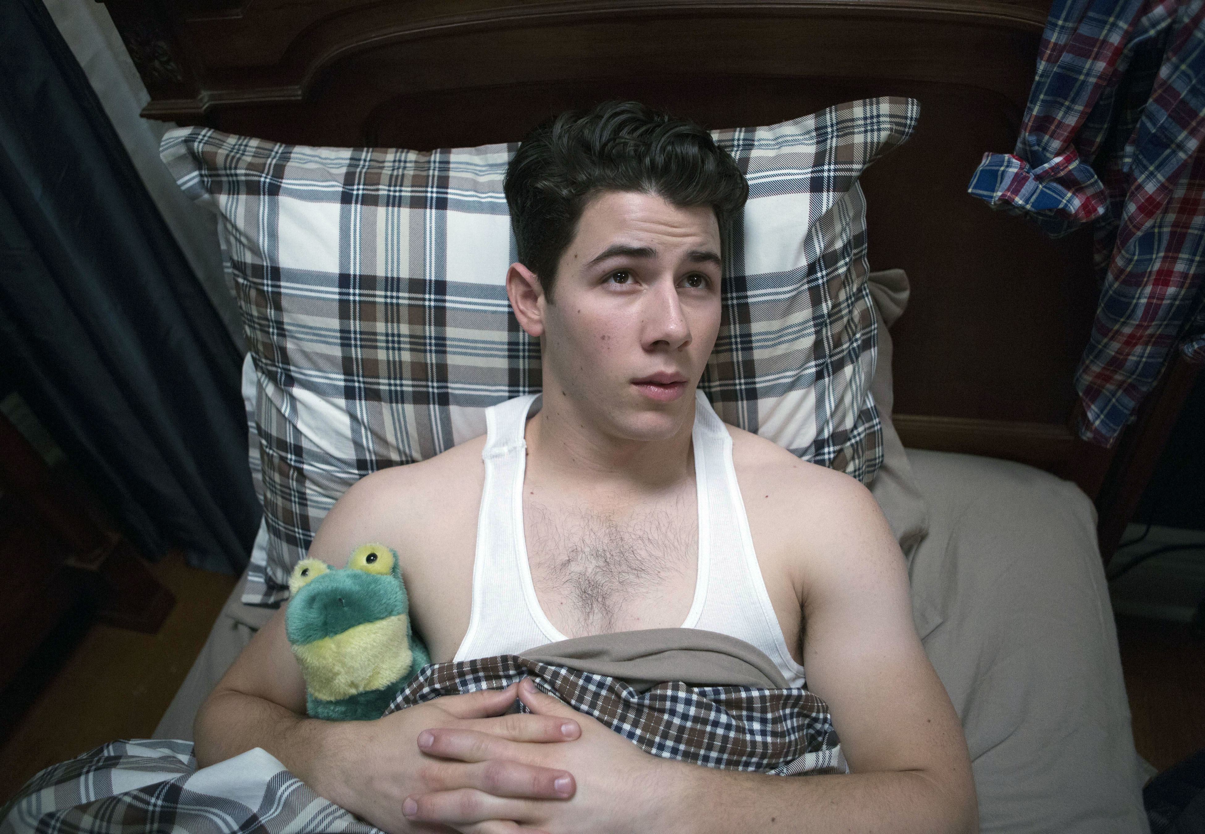 Nick Jonas lies in bed beside a stuffed frog toy, wearing a white tank top, staring upward