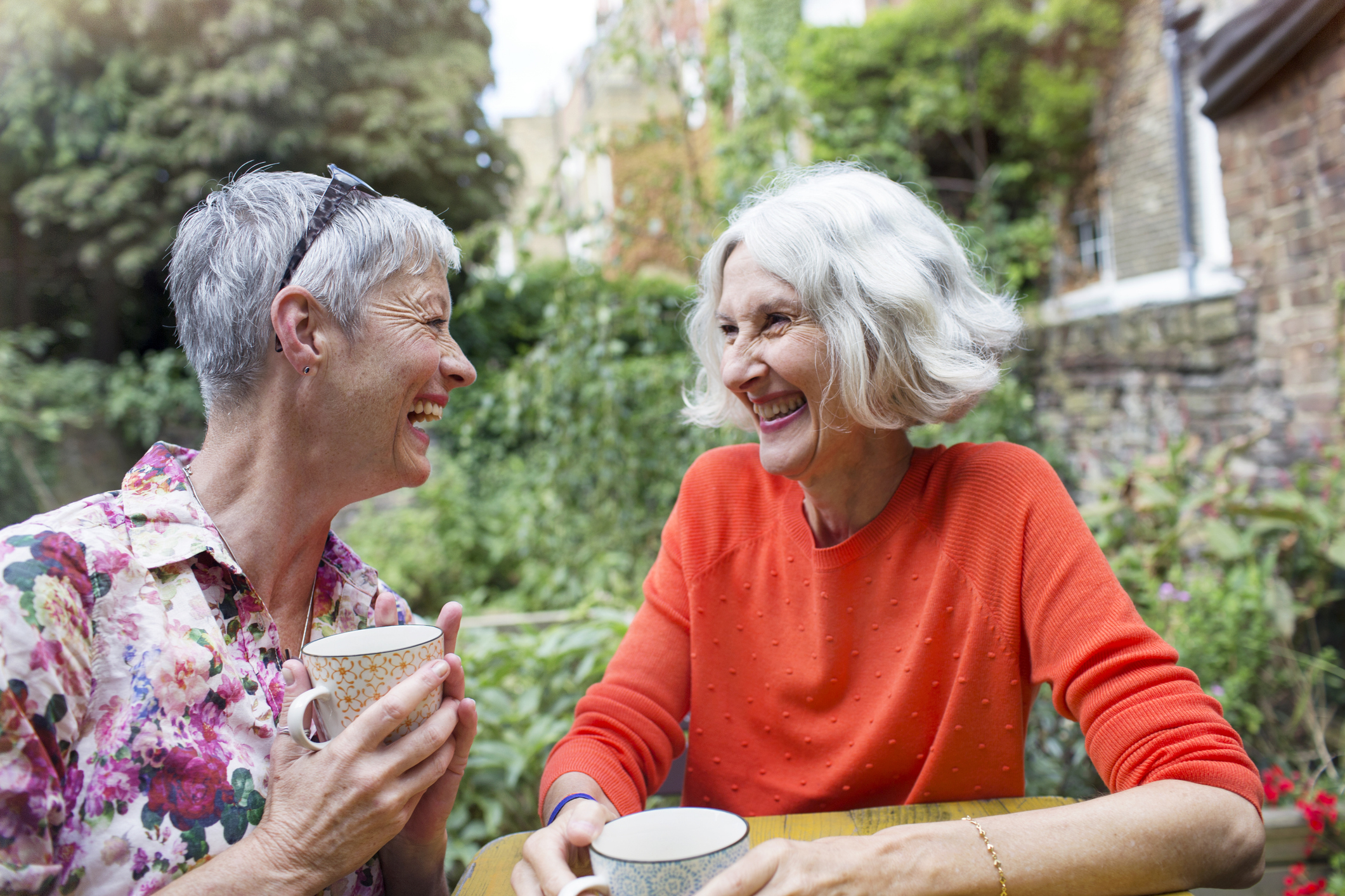 Two senior women, one in a floral shirt and the other in a red sweater, laugh together while holding teacups outdoors in a garden