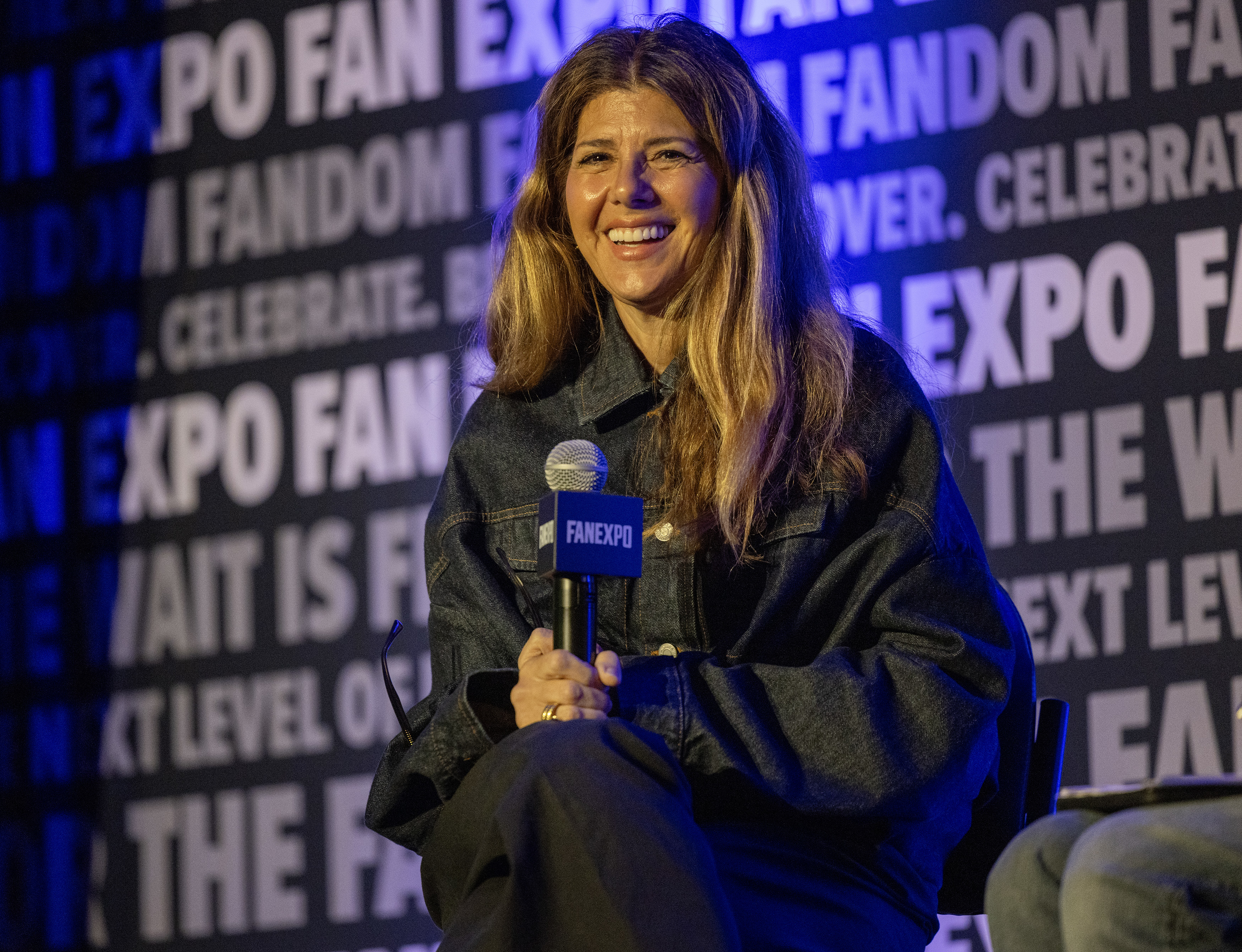 Marisa Tomei smiles while holding a FAN EXPO microphone at a panel event. She is wearing a dark denim jacket