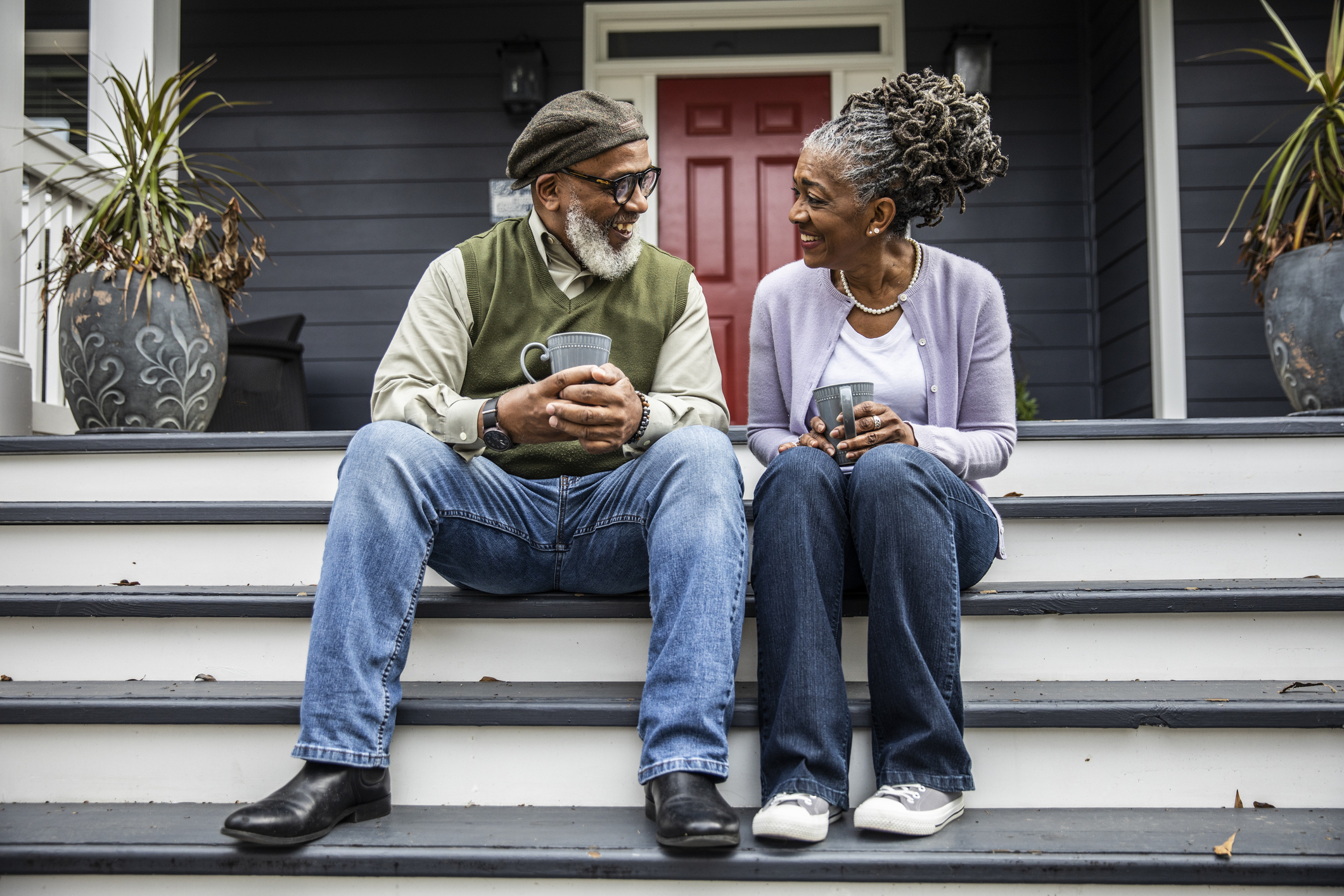 Older couple sitting on porch steps, both smiling and holding mugs. The man wears a hat, glasses, and casual clothing; the woman has gray dreadlocks and casual clothing