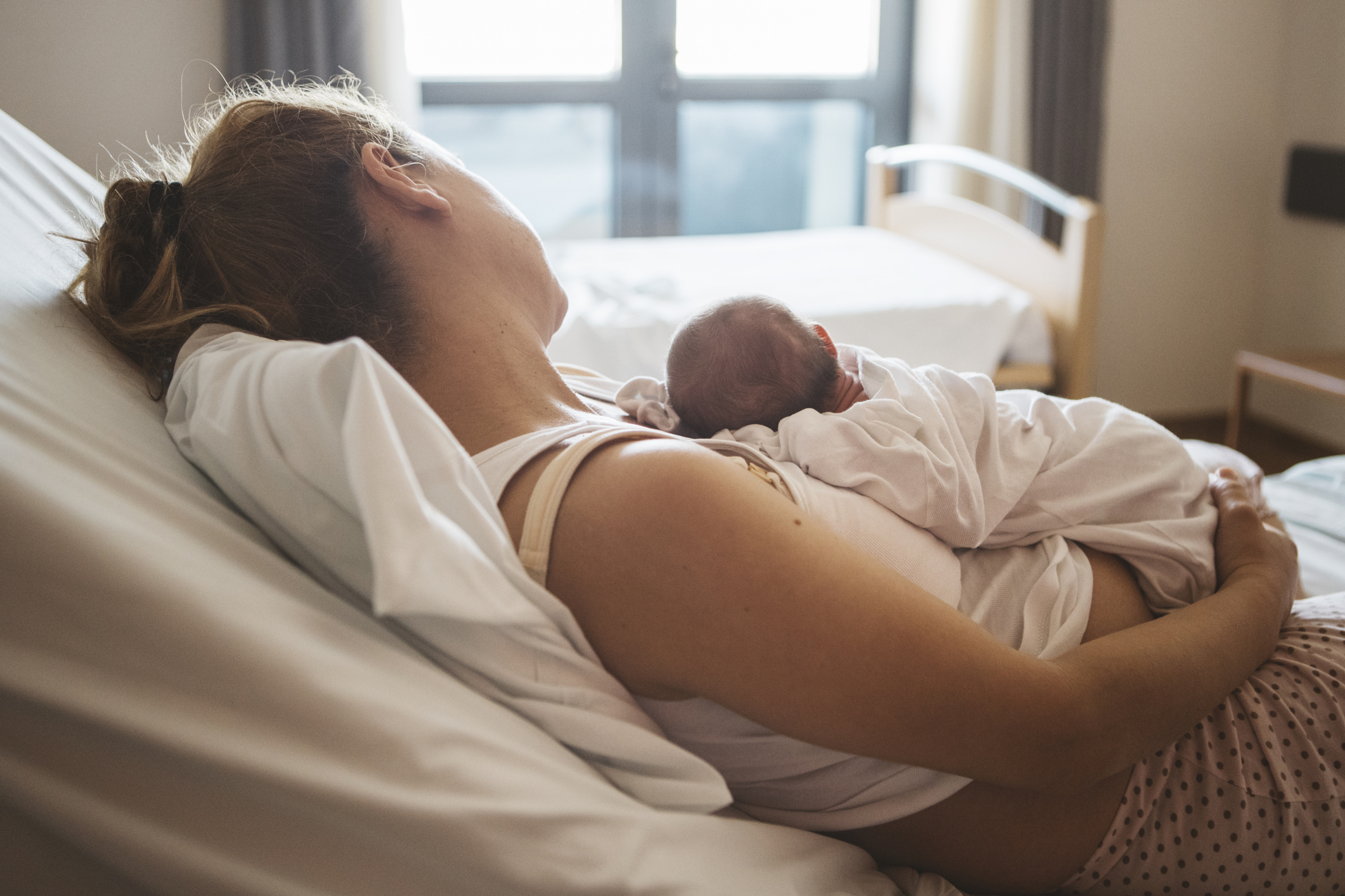 A woman lies in a hospital bed, resting with a newborn baby on her chest. The room is softly lit and appears quiet