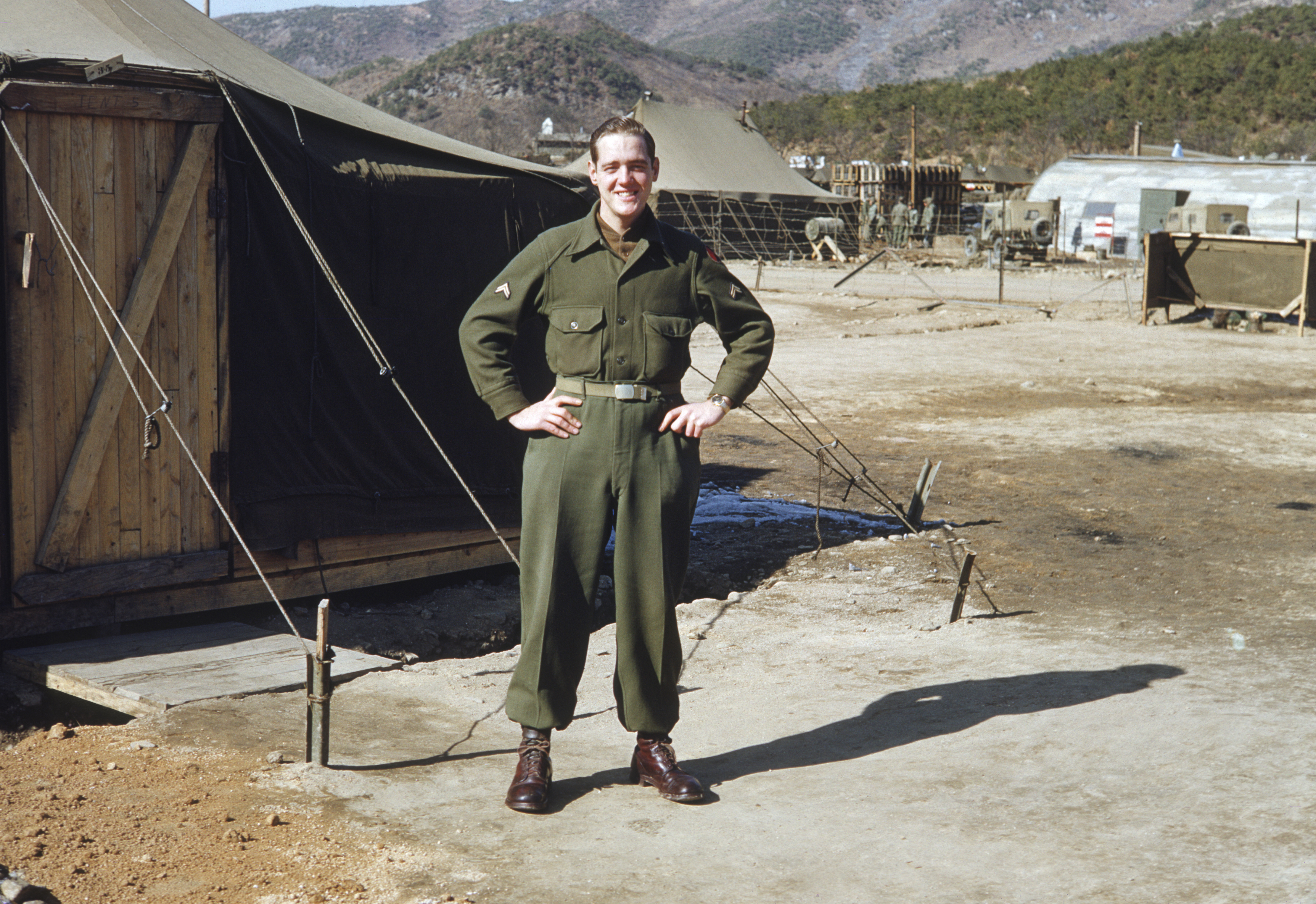 A person in military uniform stands smiling outside a tent in a camp with mountains in the background