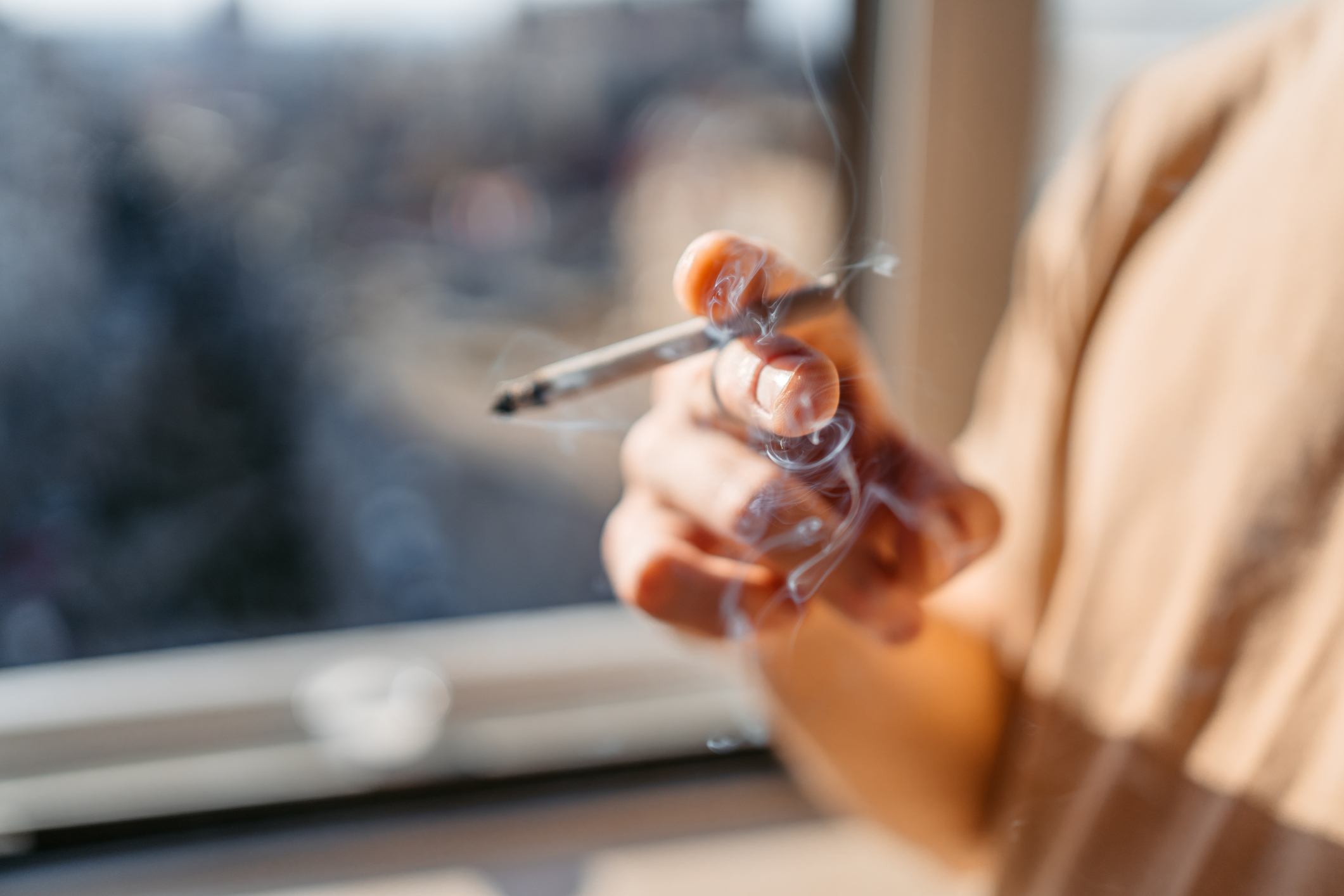 A hand holding a lit cigarette with smoke rising, set against a blurred background of a window. The person holding the cigarette is not fully visible