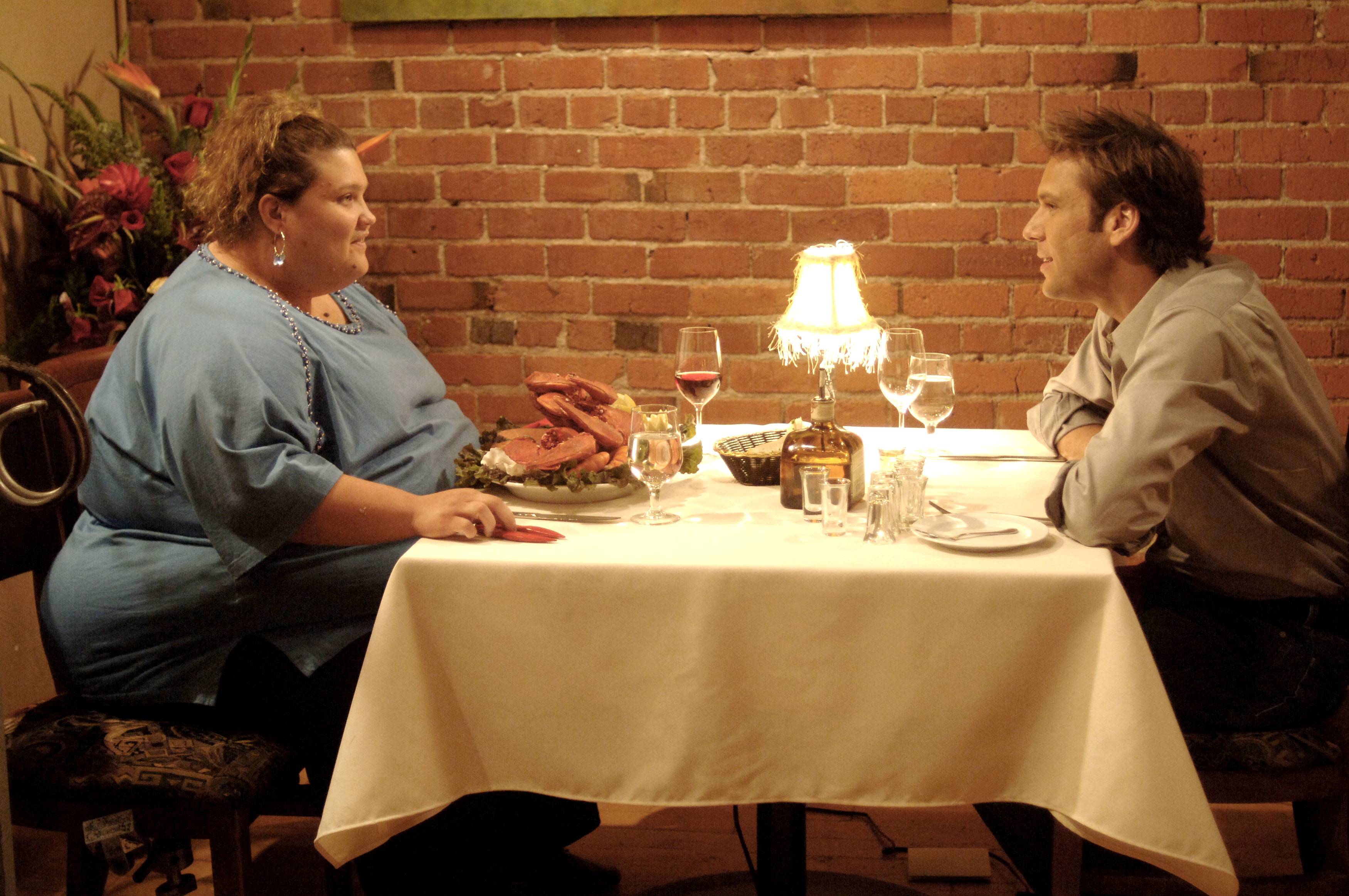 Two people, one in a blue dress and one in a blue shirt, are seated across a table with food, wine, and a lamp on a dinner date in a brick-walled restaurant