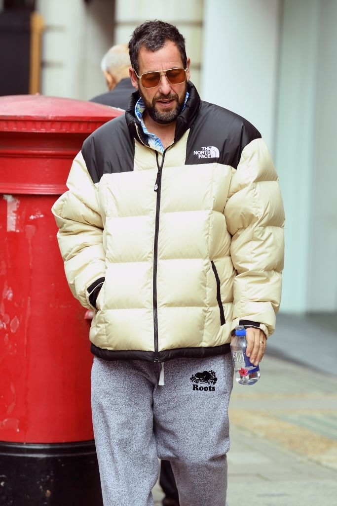 Adam Sandler in a puffy jacket and sweatpants, holding a bottle of water, walking outdoors