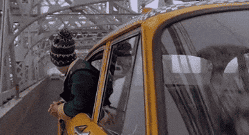 A person wearing a knit hat leans out of a moving yellow taxi, traveling on a bridge