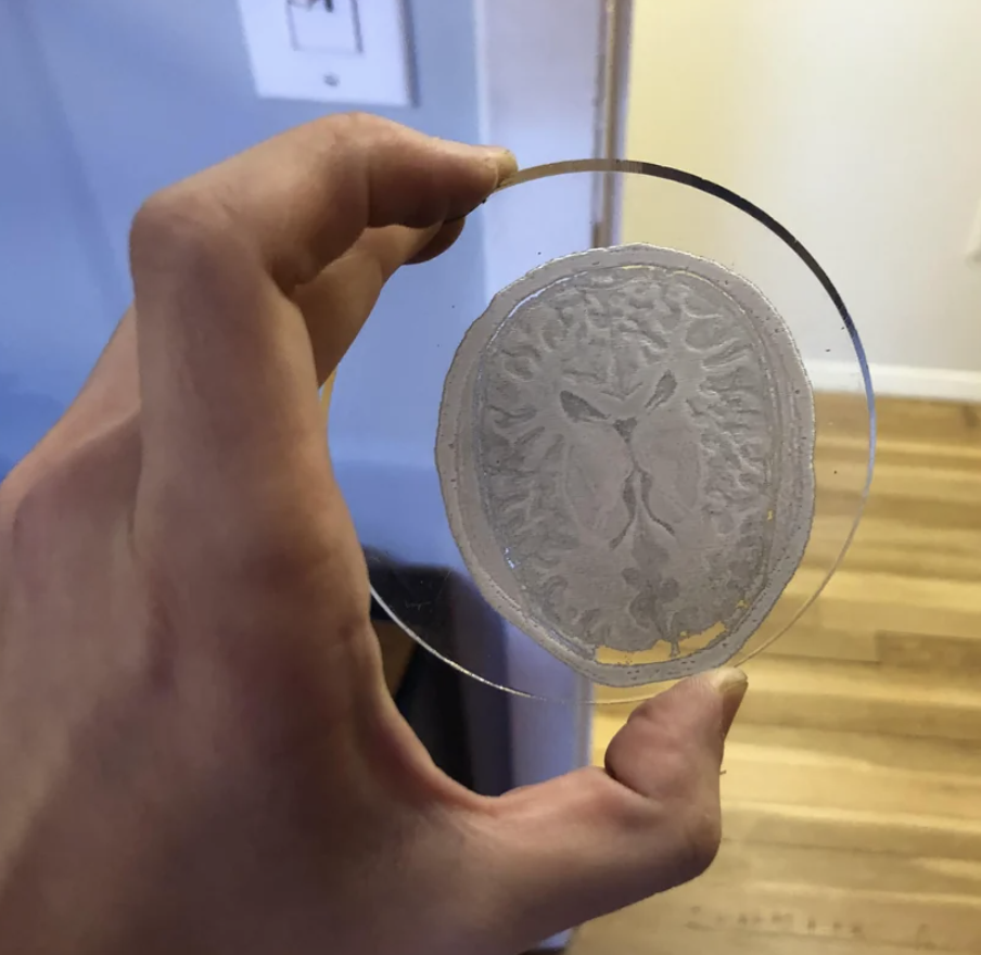 A hand holding a circular glass piece with an etched design resembling a brain scan. Background shows an interior space with wood flooring