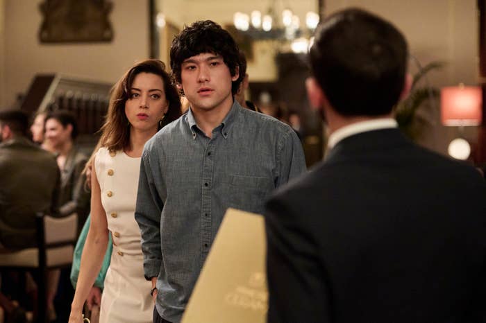Aubrey Plaza in a sleeveless dress and Jeff Baena, both looking serious, stand behind a man holding a menu at a restaurant