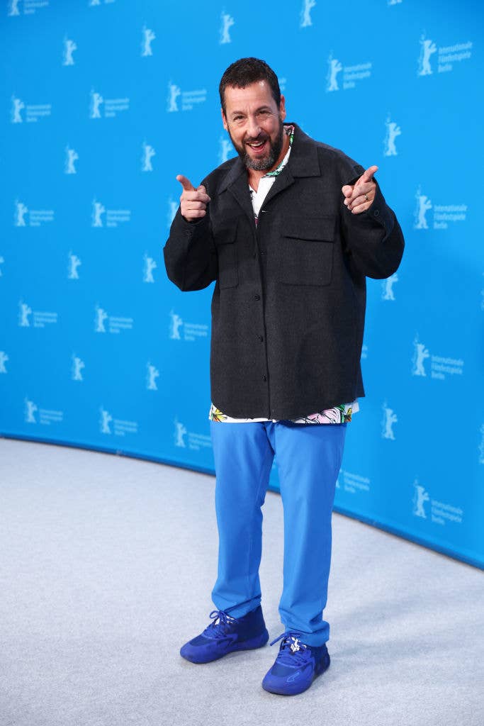 Adam Sandler smiles and points while posing at a press event. He's wearing a casual jacket, a patterned shirt, and loose-fit pants with sneakers