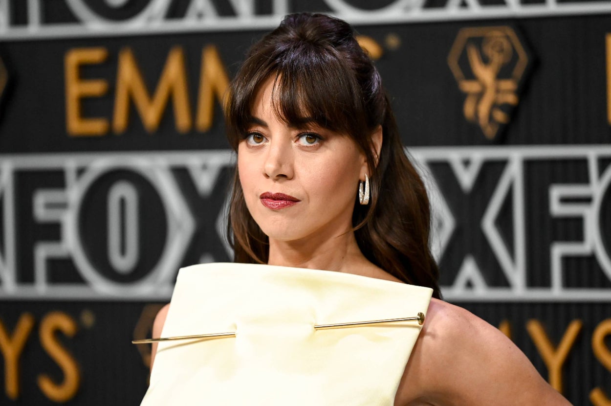 Aubrey Plaza in a stylish sleeveless dress with a unique square neckline, large hoop earrings, and bangs at the Emmys event with a backdrop featuring the FOX logo