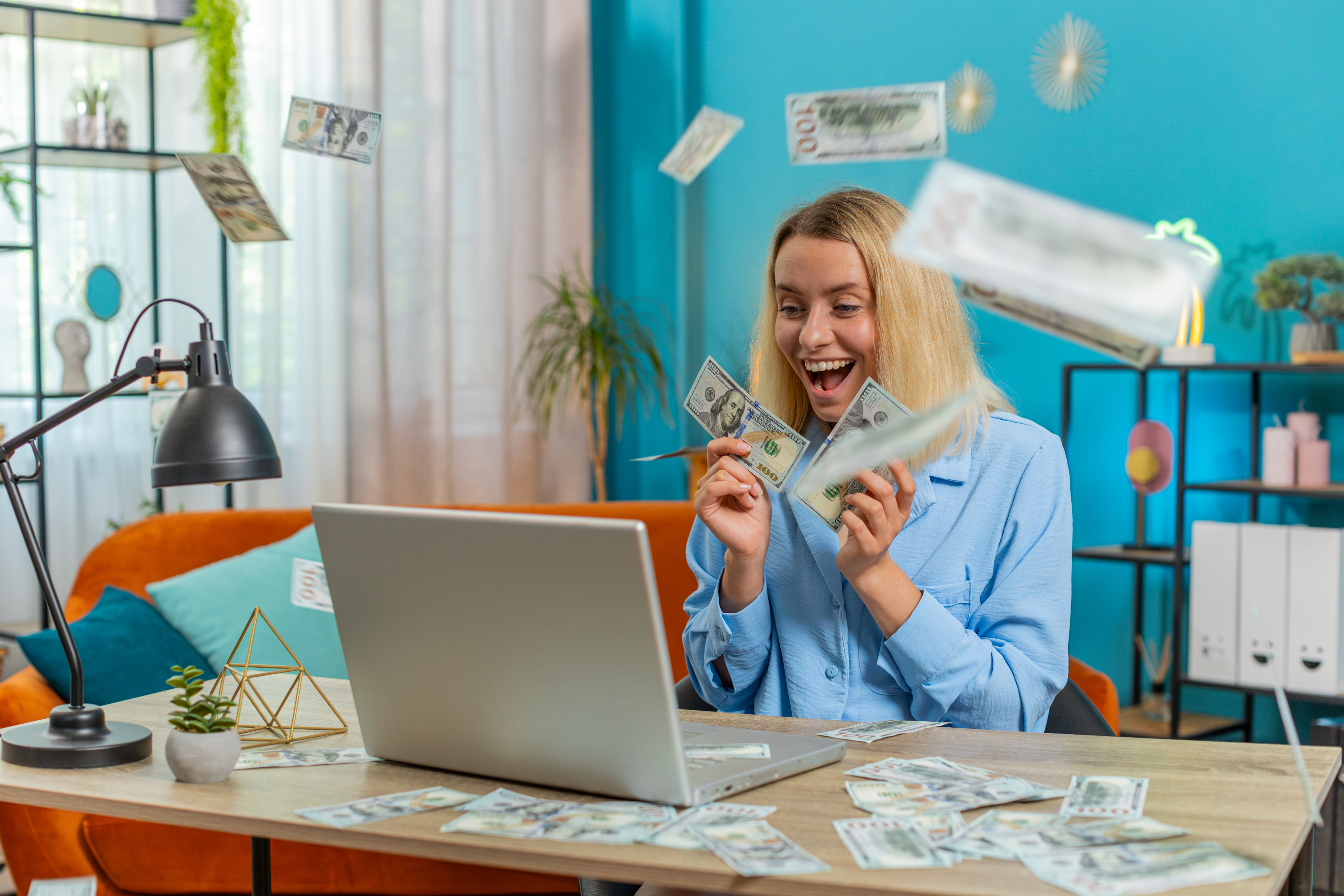 A woman smiles excitedly while holding dollar bills in front of a laptop, with more dollar bills falling around her in a lively workspace