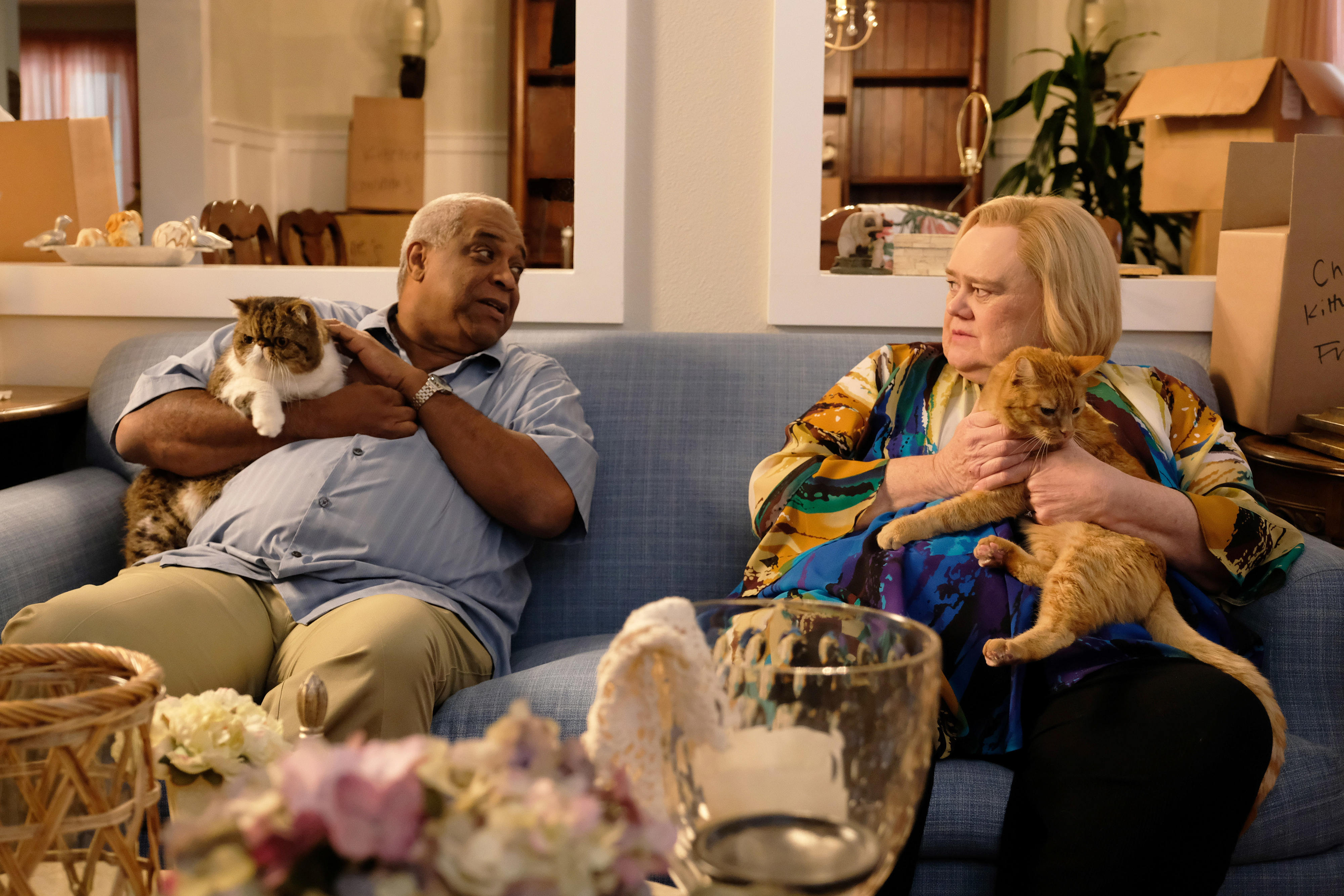 Ron Cephas Jones and Laurie Metcalf sit on a couch, each holding a cat, surrounded by moving boxes