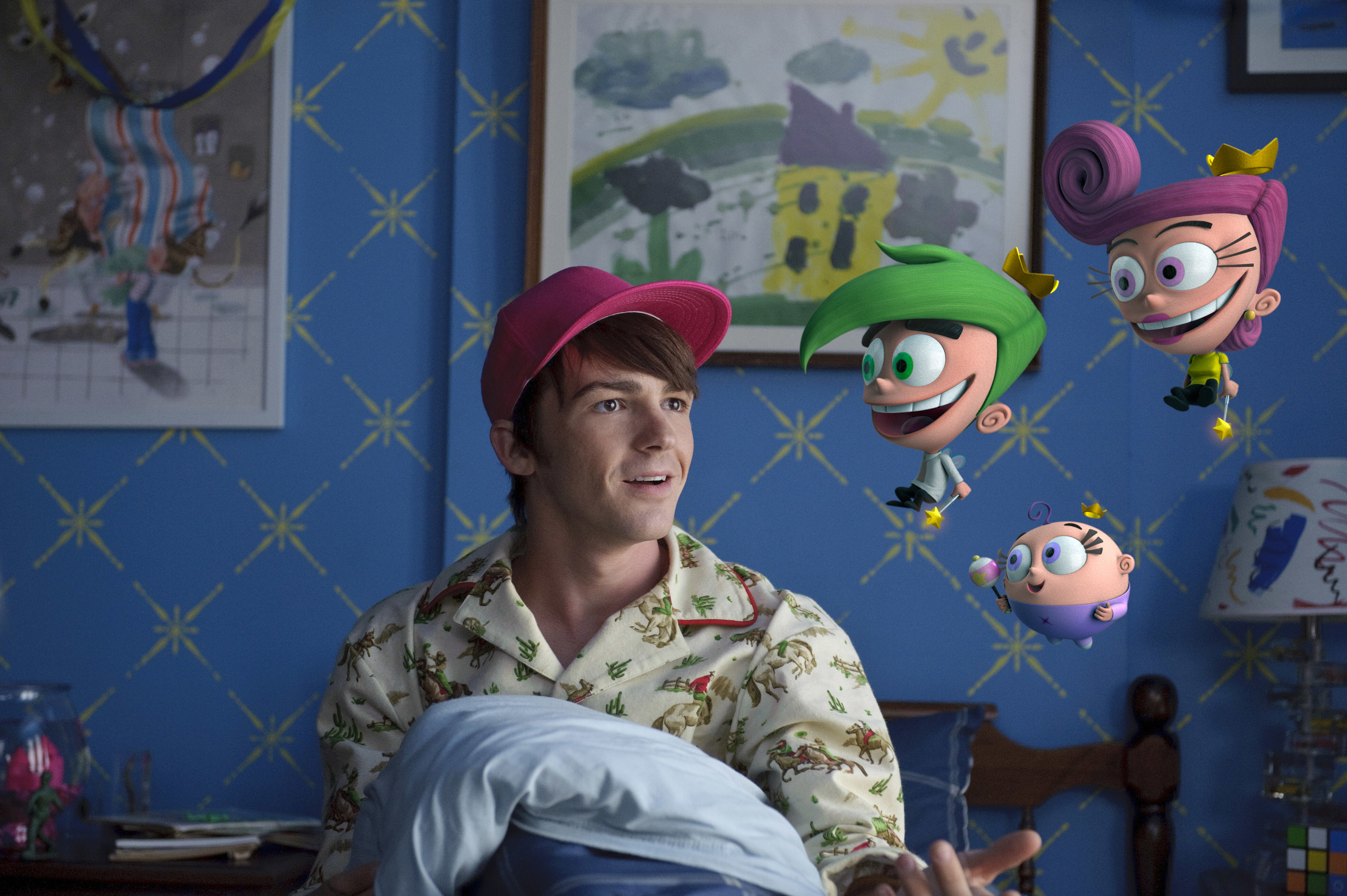 Drake Bell sits on a bed, holding a pillow, with animated characters Cosmo, Wanda, and Poof from "The Fairly OddParents" floating around him