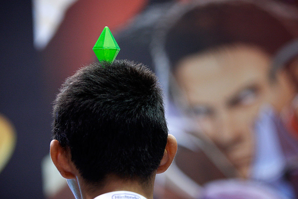 A person with short hair is seen from behind, wearing a green plumbob from The Sims on their head, with a blurred background of a face