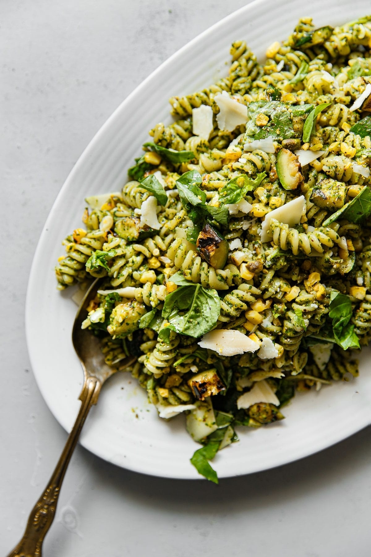 A plate of fusilli pasta salad with zucchini, spinach, nuts, parmesan cheese, and pesto sauce, garnished with fresh herbs and served with a silver fork