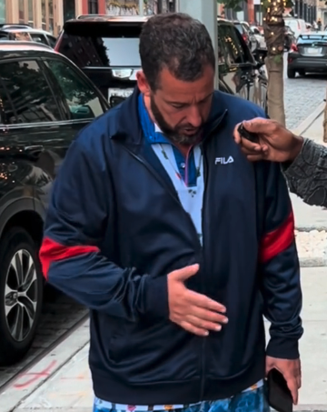 Man in a Fila jacket looks down at his hand while someone touches his shoulder on a street with parked cars in the background