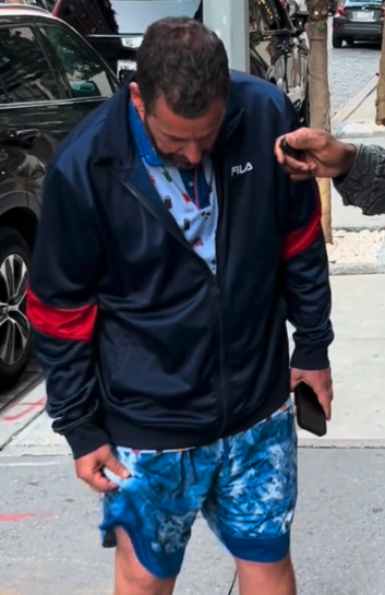 Adam Sandler wearing a track jacket and patterned shorts, looking down while standing on a sidewalk