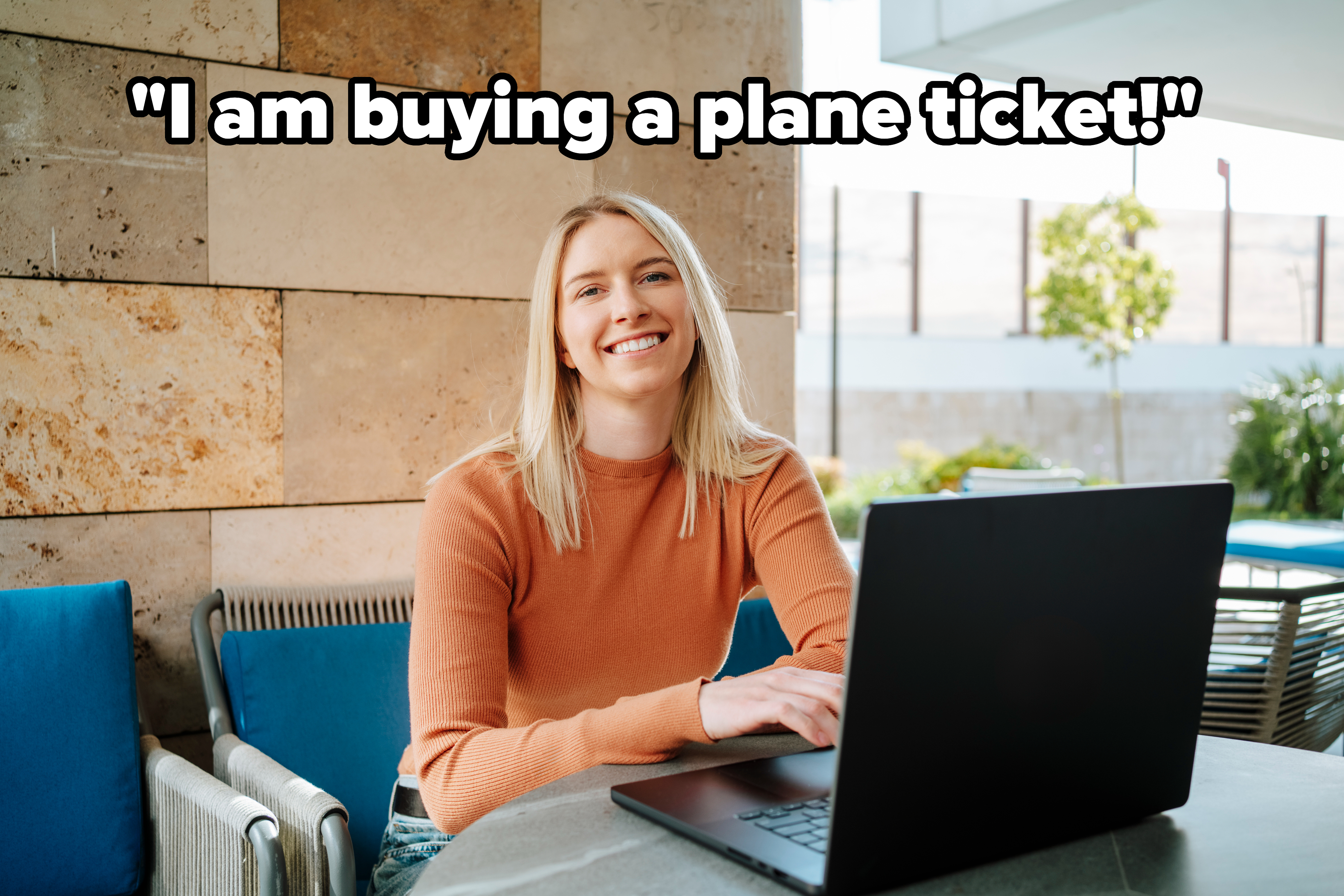 A woman in a long-sleeve top is smiling while sitting at an outdoor table with a laptop