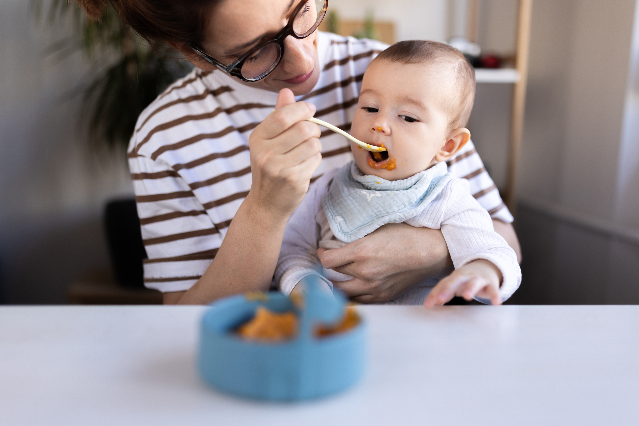 A person wearing glasses feeds a baby at a table with a small blue bowl in the foreground
