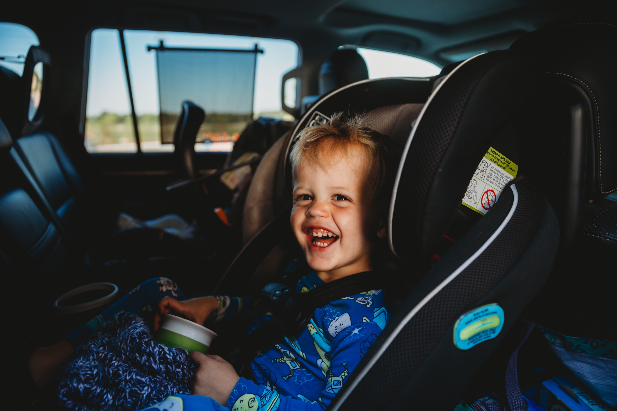 Young child smiling while seated in a car seat, holding a small cup, inside a vehicle