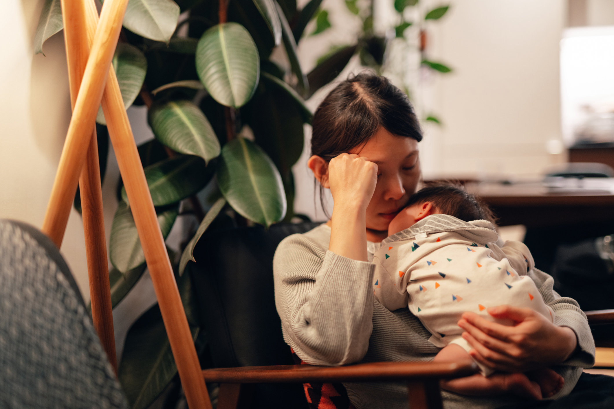 A woman sits cuddling a baby in a peaceful room with large green plants in the background