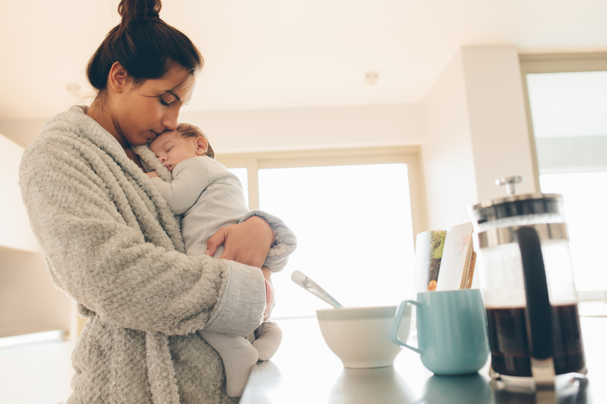 A woman in a cozy robe gently holds a sleeping baby in a kitchen with a bowl, cup, and French press on the counter